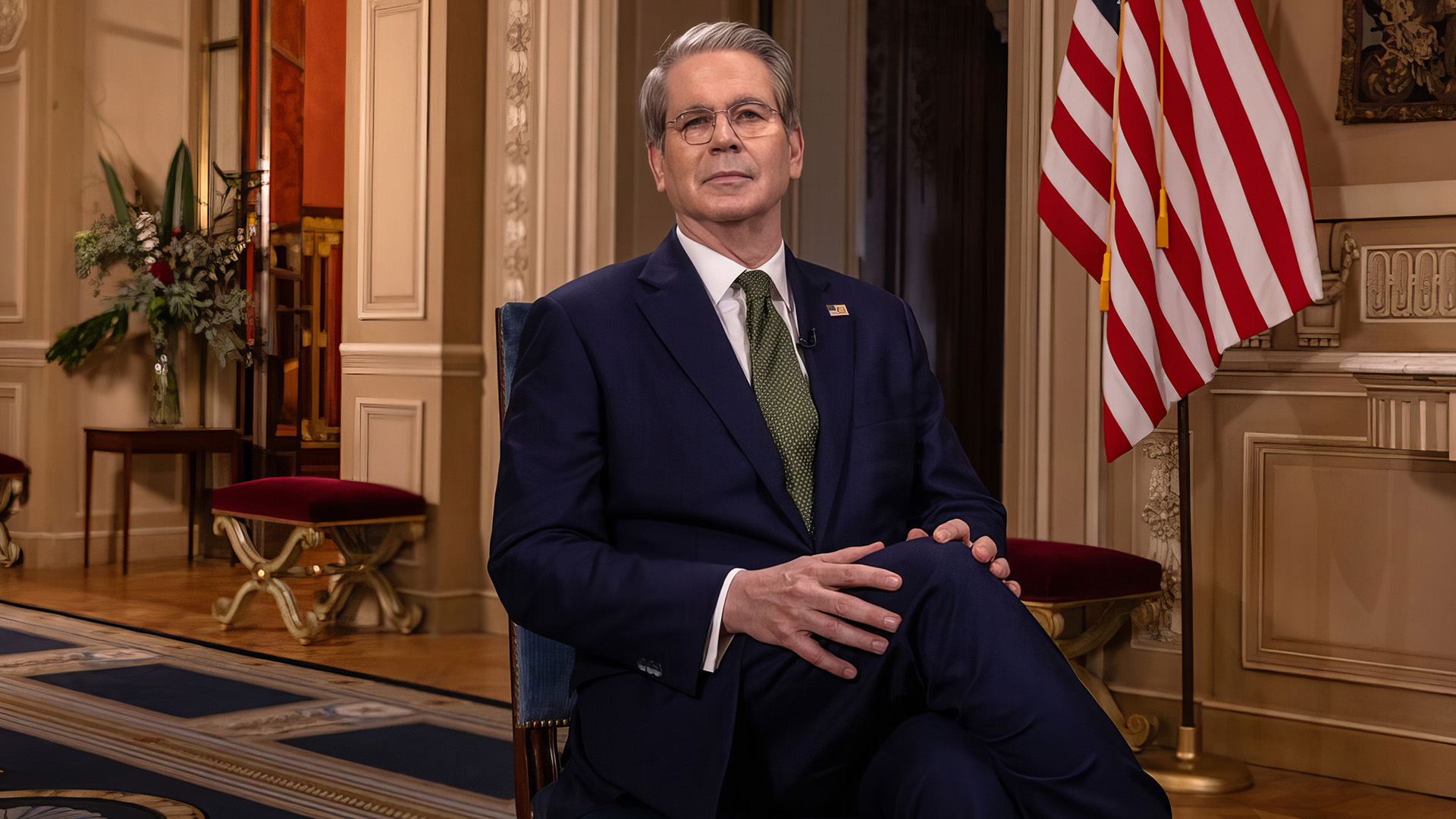 Scott bessent in a dark blue suit and green tie sits crosslegged in a formal buenos aires interview setting with an american flag visible behind him